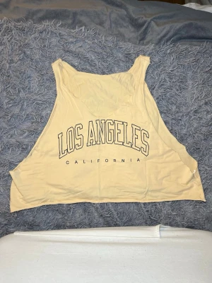 Beige topp med Los Angeles-tryck - Säljer en beige topp med trycket 'Los Angeles California' i svart på framsidan. Toppen är ärmlös och har en loose passform med breda axelpartier. Materialet är mjuk bomull (94% bomull och 6% Elastane), perfekt för varma dagar och chill stunder. Jag har klippt toppen själv. Det står ingen storlek, men skulle säga XL
