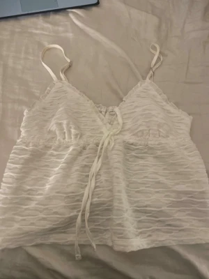 Vit omlott linnetopp med snörning - Säljer en vit omlott linnetopp med smala axelband och snygg snörning framtill. Toppen har ett vågigt, transparent mönster i tyget som ger en cool struktur. Perfekt för varma dagar och kan stylas med jeans eller kjol.