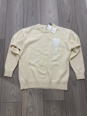 Beige stickad tröja från Ami - Säljer en beige stickad tröja från Ami med långärmad passform och ribbade muddar. Tröjan har ett vitt hjärta och bokstaven A broderat på bröstet. Perfekt för dig som gillar stilrena och mjuka plagg med snygga detaljer.