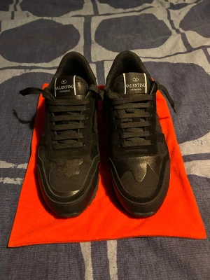 Svarta Valentino Garavani Rockrunners  - Säljer ett par svarta Valentino Garavani sneakers med snygga detaljer och mönster på ovansidan. Skorna har snörning, Materialet är en mix av skinn och syntet, vilket ger en cool och modern look. Perfekta för dig som gillar exklusiva märken och stilrena färger. Har använt dessa endast 2 gånger ny pris ungefär 6000kr