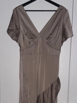 Champagnefärgad långklänning Bubbleroom - Elegant långklänning från Bubbleroom i champagnefärgad satin med v-ringad hals och korta ärmar. Klänningen har en mjuk glans och ett stilrent snitt med lätt drapering nedtill. Perfekt för dig som gillar en klassisk och chic look.