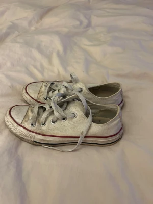 Converse All Star vita låga sneakers - Säljer ett par klassiska Converse All Star sneakers i vitt med lågt skaft. Skorna har snörning, rund tå och platt sula. De är tillverkade i bomullstyg med röda och svarta detaljer på sulan. Perfekta för dig som gillar ikonisk streetstil.