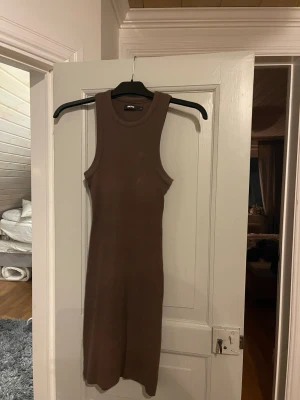 Brun ribbad ärmlös klänning från Bik Bok - Säljer en brun, ribbad klänning från Bik Bok. Modellen är ärmlös med rund hals och har en tight passform som framhäver figuren. Klänningen är av stickad bomull och slutar strax under knäna, perfekt för en enkel och stilren look.