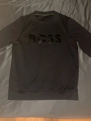 Svart tröja från BOSS - Svart långärmad sweatshirt från BOSS med stor, glansig logotyp på bröstet. Tröjan har rund hals med en diskret grön detalj på insidan av kragen. Materialet är mjukt och känns som bomull, med en ribbad struktur som ger extra stilpoäng.
