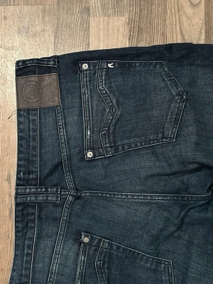 Replay jeans i mörkblå denim - Säljer ett par snygga Replay jeans i mörkblå tvätt med klassisk femficksdesign och raka ben. Jeansen har tydliga kontrastsömmar, läderpatch bak och Replay-logga vid fickan. Materialet är slitstark bomullsdenim som ger en cool och tidlös look.