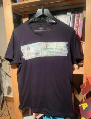 Svart t-shirt med konstnärligt tryck - Svart t-shirt från South West i bomull med korta ärmar. På bröstet finns ett konstnärligt tryck i vitt, blått och grönt med handritade blad och linjer, vilket ger en unik och kreativ vibe. Perfekt för dig som gillar personliga detaljer och grafiska motiv.