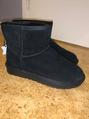 Svarta mockaskor - Säljer ett par svarta uggs i mocka med rund tå och platt sula. Skorna har ett mjukt och fluffigt foder på insidan och är tillverkade i skinn. Perfekta för kalla dagar och har en enkel, stilren design utan synliga detaljer eller mönster.