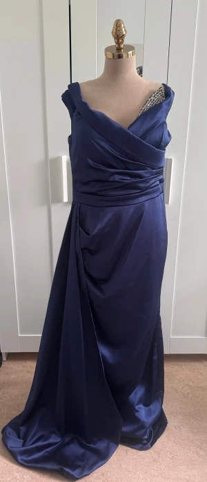 Marinblå långklänning från Zeropoint - Säljer en marinblå långklänning från Zeropoint med offshoulder-skärning och draperad överdel. Klänningen har en elegant omlott-look och är tillverkad i ett glansigt, mjukt polyestertyg. Perfekt för dig som vill sticka ut med en klassisk och stilren look.
