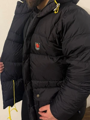 Svart Fjällräven Polar dunjacka S - Säljer en svart dunjacka från Fjällräven Polar med huva och gula snören. Jackan har flera fickor med knappar, broderad logga på bröstet och dragkedja samt tryckknappar framtill. Perfekt för kalla dagar och riktigt snygg streetstil.