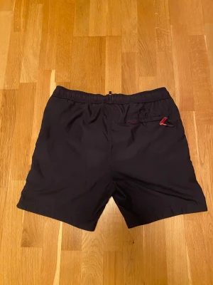 Svart badväst Superdry med tryck - Svart badshorts från Superdry i storlek M med vitt tryck '09' på framsidan och röda snören i midjan. Shortsen har en bakficka med röd dragkedja och insida i rött tyg. Perfekt för strand eller poolhäng.