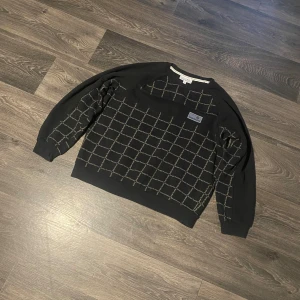 Svart sweatshirt från Sergio Tacchini - Svart sweatshirt från Sergio Tacchini med rutigt mönster och logotyp på bröstet. Tröjan har rund hals, ribbade muddar och lång ärm. Materialet är mjuk bomull och polyester, perfekt för chill dagar. Snyggt rutmönster med diskret branding över hela framsidan.