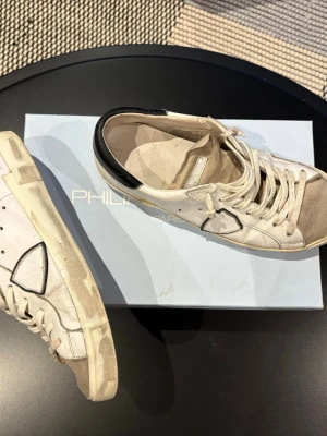 Philippe Model vita sneakers med svart häl - Säljer ett par vita Philippe Model sneakers med svart detalj på hälen och bruna gummisulor. Skorna har snörning och en rund tå, samt en ikonisk sköldpatch på sidan. Materialet är skinn och de har en klassisk platt sula. Perfekt för dig som gillar fransk streetstyle. Dock är ena sulan lite fuckt men därför priset går säkert att lima elle nått 