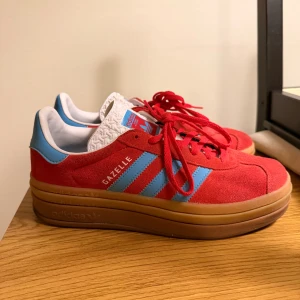 Adidas Gazelle röda sneakers med platåsula - Säljer ett par Adidas Gazelle sneakers i rött mocka med ljusblå ränder och detaljer. Skorna har en chunky brun platåsula, klassisk snörning och vit plös med präglat mönster. 