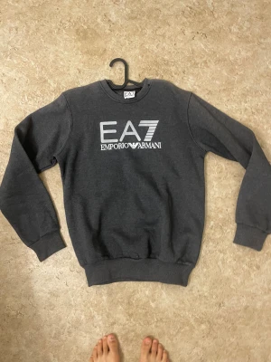 Mörkgrå EA7 Emporio Armani sweatshirt - Snygg mörkgrå sweatshirt från EA7 Emporio Armani med stort vitt tryck på bröstet. Tröjan har rund hals, ribbade muddar vid ärmslut och nederkant samt långärmad passform. Perfekt för dig som gillar stilrena märkesplagg.
