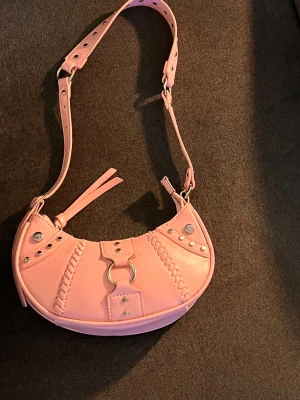 Rosa axelväska i skinn med nitar - Snygg rosa axelväska i skinn med justerbart axelband, silverfärgade nitar och dekorativa sömmar. Väskan har dragkedja upptill och coola metalldetaljer framtill. Perfekt för dig som vill sticka ut med en trendig accessoar.