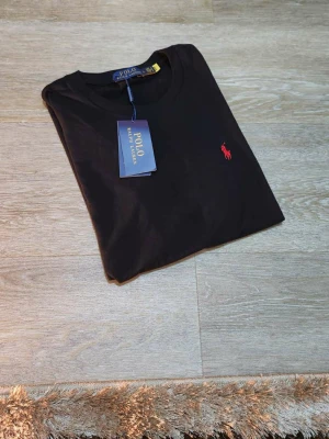 Svart t-shirt från Polo Ralph Lauren - Svart t-shirt från Polo Ralph Lauren med klassisk rund hals och korta ärmar. T-shirten har det ikoniska röda broderade polospelar-loggan på bröstet. Tillverkad i mjuk bomull för en skön känsla och clean look.