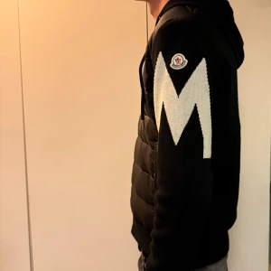 Svart Moncler pufferjacka med huva - Storlek XL men passar L. Svart pufferjacka från Moncler med huva och dragkedja framtill. Jackan har stickade ärmar med en stor vit M-detalj på vänster arm och Moncler-logga. Kroppen är quiltad och har snörning vid huvan. Snygg och modern design som sticker ut.