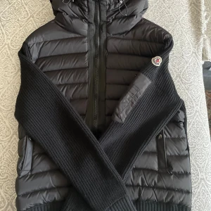 Svart Moncler pufferjacka med stickade ärmar - Svart pufferjacka från Moncler med stickade ärmar och rygg, quiltad framsida och huva. Jackan har dragkedja framtill, fickor med tryckknappar och en extra ficka med dragkedja på ärmen. Klassisk Moncler-logga på vänster ärm. Perfekt för kyliga dagar.