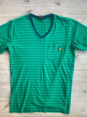 Grön randig t-shirt från Lyle & Scott - Snygg grön t-shirt från Lyle & Scott med tunna mörkblå ränder och v-ringad hals. T-shirten har en bröstficka med den klassiska gula fågel-loggan. Kortärmad och tillverkad i mjuk bomull, perfekt för en chill stil.