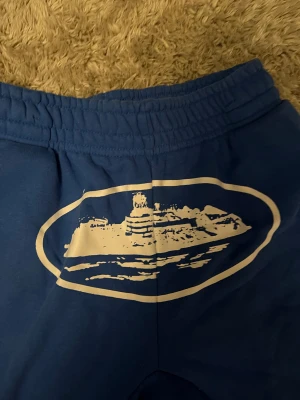 Blåa corteiz alcatraz shorts - Blå mjukisbyxor i kort modell med resår i midjan och ett stort vitt tryck framtill som föreställer en byggnad. Byxorna har fickor på sidorna och en bakficka med knapp. Mjukt material som känns skönt mot huden.