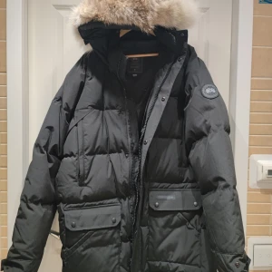 Svart dunjacka från Canada Goose - Säljer en svart dunjacka från Canada Goose med fluffig päls på huvan. Jackan har flera stora fickor med knappar, dragkedja och en klassisk patch på ärmen. Perfekt för kalla vinterdagar och har en riktigt schysst oversized look.