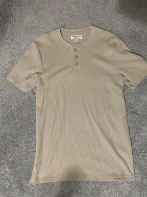 Beige ribbad t-shirt från Denim Day - Säljer en beige ribbad t-shirt från Denim Day i storlek small. T-shirten har korta ärmar, rund hals och tre knappar framtill. Materialet är mjukt och känns som bomull, perfekt för en chill och enkel stil.