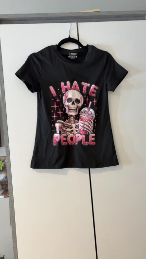 Svart t-shirt med skelettmotiv XS - Svart t-shirt i polyester med tryck av ett skelett som håller en milkshake och texten 'I HATE PEOPLE' i rosa och vitt. T-shirten har korta ärmar och rund hals. Perfekt för dig som gillar edgy och humoristiska prints.