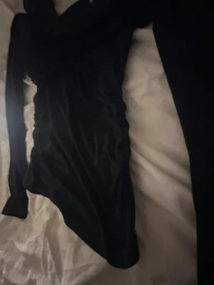 Svart omlott topp från Zara - Svart omlott topp från Zara med lång ärm och snygg rynkning längs sidorna. Modellen är figurnära och har en cool asymmetrisk skärning vid midjan. Perfekt för dig som gillar stilrena och trendiga plagg med lite edge.