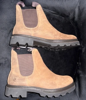 ECCO – Chelsea boots  - Stilrena chelsea boots i mjuk mocka från ECCO. Robust sula och ren silhuett – byggda för vardag, stad och säsongsskifte.  Skick: Nya Storlek: 38  Färg: Brun Material: Mocka Nypris: ca 2 000 kr The Label Drop-pris: 1 000 kr  The Label Drop – Premium kvalitet för alla. 