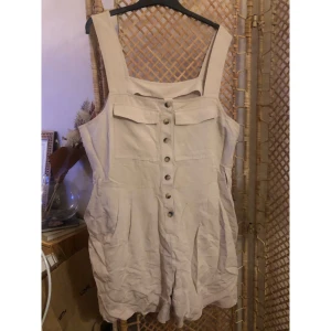 Beige byxdress från H&M, storlek 50 - Snygg beige byxdress från H&M i storlek 50. Modellen har breda axelband, knappar framtill och två bröstfickor. Tillverkad i ett lätt och luftigt material, perfekt för varmare dagar. Stilren och enkel att matcha med olika accessoarer. Denna är mer som en large i storlek. 