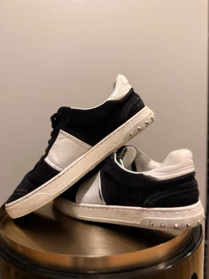 Valentino Garavani svartvita sneakers - Svartvita Valentino Garavani sneakers med klassisk låg siluett. Skorna har svart mocka och vitt skinn, svarta snören och coola nitar på hälen. Perfekt för dig som gillar stilrena men ändå edgy detaljer. 17/21 nitar kvar. Stor i storlek så passar 42 också.