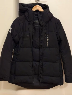 Svart dunjacka Sail Racing med huva - Säljer en svart dunjacka från Sail Racing med broderade loggor på bröstet och ärmarna. Jackan har en rejäl huva, dragkedja framtill och flera praktiska fickor. Perfekt för kalla dagar och har en clean, sportig look.