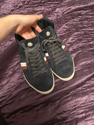 Moncler svarta mocka vinterskor - Snygga svarta sneakers från Moncler i mocka med högt skaft. Skorna har vita och röda detaljer på sidan med Moncler-logga, bruna skosnören och vit sula. Klassisk rund tå och stilren design som passar till många outfits. Bra nu till vintern