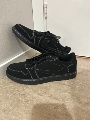 Nike Jordan 1 Low svart sneakers - Svarta Nike Jordan 1 Low sneakers med kontrasterande vita sömmar och klassisk Swoosh på sidan. Skorna har snörning, rund tå och platt sula. Materialet är syntet och skinn, vilket ger en clean och stilren vibe. Perfekt för dig som gillar streetwear och vill ha en ikonisk look.