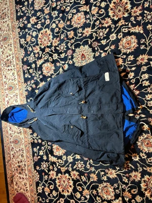 Adidas Originals marinblå parkajacka - Parkajacka från Adidas Originals i marinblått med blått foder. Jackan har huva med snörning, två stora fickor med lock och knappar samt dragsko i midjan. Materialet är polyester och jackan har en loose passform som är chill och bekväm.