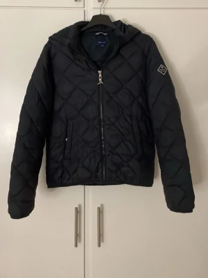 Svart quiltad jacka från Gant - Quiltad svart jacka från Gant med huva och dragkedja framtill. Jackan har ett snyggt rutmönster och en patch med Gant-logga på ärmen. Perfekt för dig som gillar stilren och sportig look. Långärmad och med två fickor framtill.