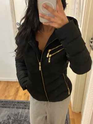 Svart pufferjacka med guldiga dragkedjor - Snygg svart pufferjacka med flera guldiga dragkedjor och huva. Jackan har figurnära passform, quiltad design och coola detaljer som ger en modern vibe. Perfekt för dig som vill ha en varm och stilren jacka med edge.