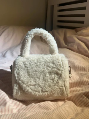 Vit fluffig handväska med axelrem - Supergullig liten handväska i vitt fuskpälsmaterial med både handtag och justerbar axelrem. Väskan har magnetlås och insida i vitt syntetmaterial med innerficka med dragkedja. Perfekt accessoar för att lyfta din outfit.