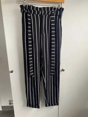 Svarta randiga byxor  - Svarta byxor med vita vertikala ränder och högt papperspåse-midja. Byxorna har ett brett knytband i midjan och raka ben. 