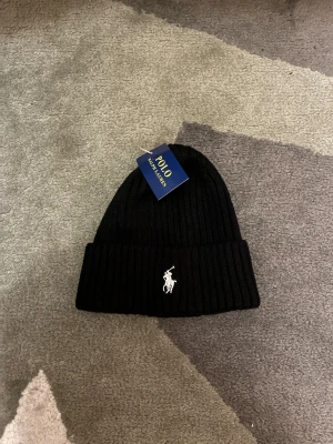 Svart Polo Ralph Lauren-mössa - En stilren, ribbstickad svart mössa med det klassiska Polo-emblemet. Perfekt för både vardag och kyliga dagar.