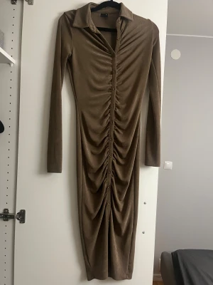 Brun långklänning Gina Tricot XS - Snygg brun långklänning från Gina Tricot i storlek XS. Klänningen har långa ärmar, krage och knappar hela vägen framtill. Snygg rynkad effekt längs mitten ger en cool och trendig look. Perfekt för dig som gillar en tight och figurnära stil.
