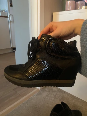 Svarta sneakers med kilklack - Säljer ett par svarta sneakers från G by Guess med kilklack och snörning. Skorna har glansiga, reptilpräglade paneler i syntetmaterial och rund tå. Snygg detalj med metallplatta på snörningen. Perfekt för dig som vill ha lite extra höjd men ändå sportig stil.