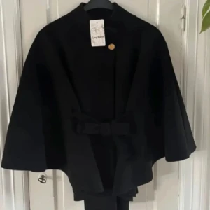Svart poncho  - Elegant svart kappa med en stilren design. Den har en knäppning framtill och ett dekorativt bälte i midjan. Perfekt för att ge en sofistikerad touch till din outfit.