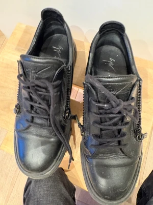 Svarta sneakers från Giuseppe Zanotti - Säljer ett par svarta sneakers från Giuseppe Zanotti med snörning och dragkedjor på sidorna. Skorna är tillverkade i skinn och har en platt sula samt rund tå. Snygg och stilren design med coola detaljer som ger extra edge till din outfit.