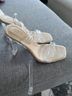 Beige sandaler med transparent klack - Säljer ett par trendiga sandaler från ASOS i beige med transparenta remmar och klack. Skorna har fyrkantig tå och en cool, genomskinlig plattformklack som ger en modern vibe. Perfekt för dig som gillar stilrena detaljer och vill sticka ut.