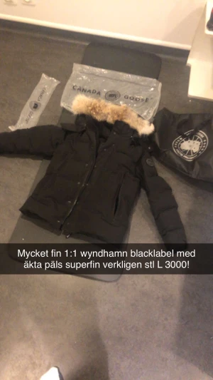 Svart Canada Goose Wyndham dunjacka L - Canada Goose Wyndham Black Label dunjacka i svart med avtagbar äkta päls på huvan. Jackan har klassisk patch på ärmen, flera fickor, dragkedja och knappar framtill. Perfekt för kalla dagar med sin tjocka och puffiga design.