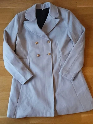 Ljusgrå kappa - Snygg ljusgrå kappa med dubbelknäppning och stora guldknappar. Jackan har klassisk krage, två rejäla fickor och är långärmad. Perfekt för kyliga dagar. Är något liten i storleken. Har aldrig blivit använd och säljes då den är för liten för mig.