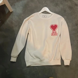 Vit sweatshirt från Ami med rött hjärta - Säljer en clean vit sweatshirt från Ami Alexandre Mattiussi med ett stort rött hjärta och bokstaven A broderat på bröstet. Tröjan har rund hals, ribbade muddar och lång ärm. Perfekt för dig som gillar stilrena detaljer och vill sticka ut lite. Pris kan diskuteras.