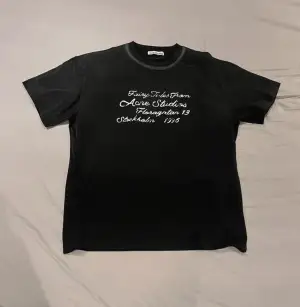 Svart t-shirt från Acne Studios med vit text på bröstet: 'Fairy Tales From Acne Studios Floragatan 13 Stockholm 1996'. Klassisk rund hals och korta ärmar. Mjuk bomullskvalitet och normal passform. Perfekt statement-plagg för dig som gillar stilrena designer.