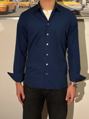 Italiensk dress shirt - Snygg mörkblå skjorta med klassisk krage och långa ärmar. Skjortan har en bröstficka och silverfärgade knappar framtill. Passformen är normal och materialet känns lätt och bekvämt, troligen bomull. Perfekt för dig som gillar stilrena och enkla plagg.
